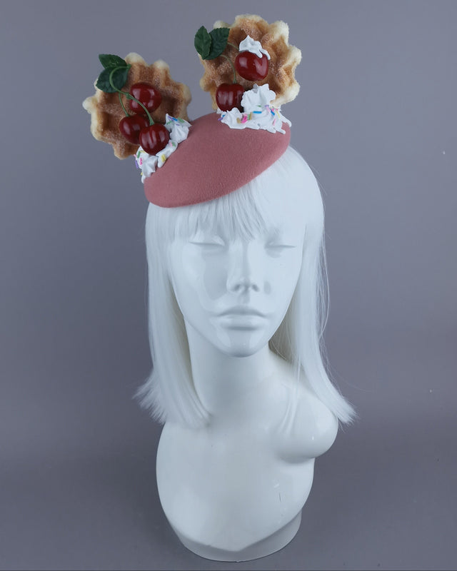 A food themed fascinator hat, set on a pink base and cream swirl on top, decorated with cherries and cream waffle ear