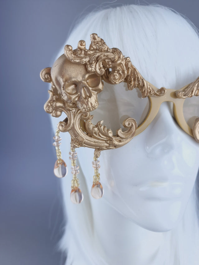 Close up of a pair of ornate gold cat-eye sunglasses with filigree, skull, and beading details displayed on a mannequin head.