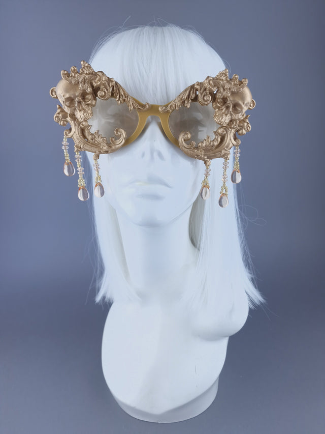 A pair of ornate gold cat-eye sunglasses with filigree, skull, and beading details displayed on a mannequin head.