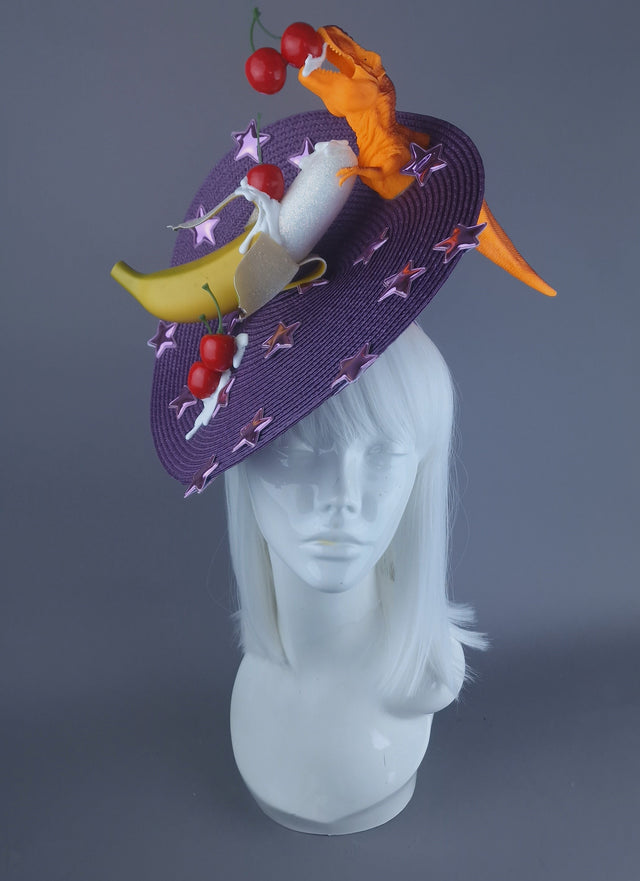 A colourful fruit and dinosaur fascinator hat featuring a purple base with metallic stars and a tyrannosaurus rex silhouette, alongside banana and cherry decorations.