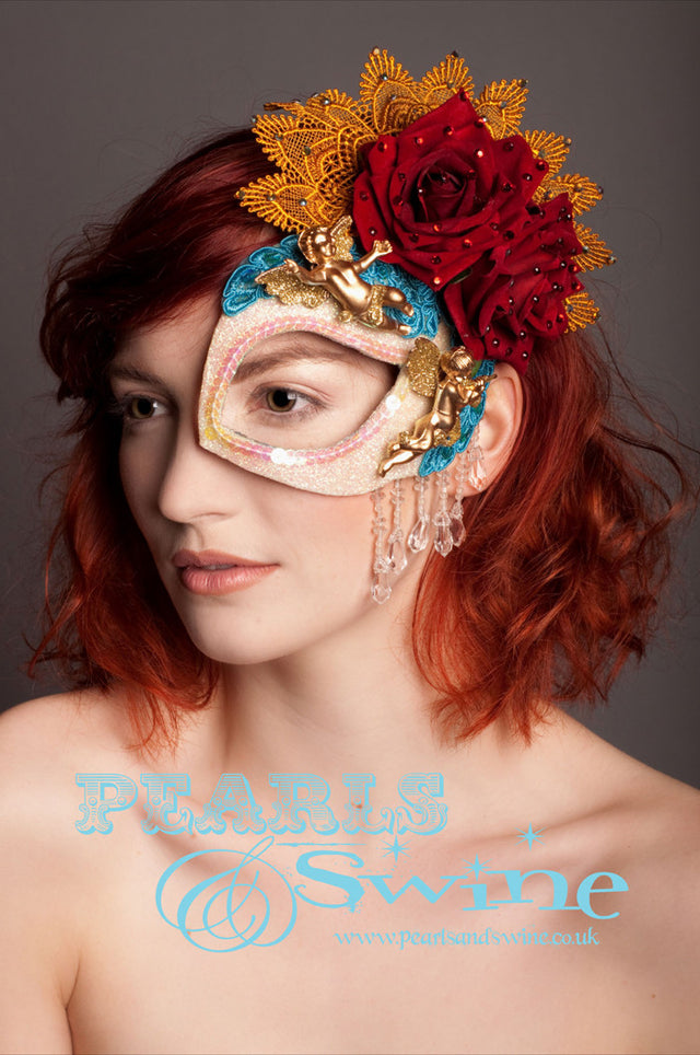 Virgin Mary Half Mask "Return To Innocence" A Virgin Mary-inspired white glitter covered half mask fascinator complete with angels, lace, crystals, scarlet velvet roses, golden cherubs. The eye is edged in clear iridescent sequins and clear fringe beading. Backed with leopard print satin and attaches to the hair with a comb and adjustable hat elastic