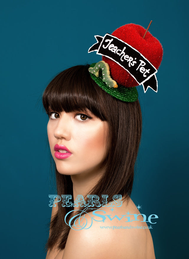 Giant red apple fascinator set on a green glitter fascinator base, decorated with a chalk board banner with the words "Teacher's Pet" and a worm. The base of our giant red apple fascinator is backed with leopard print and attaches with a comb and hat elastic. Perfect for a school girl fancy dress outfit or burlesque performer.