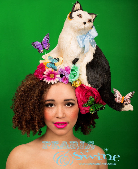 Surreal Cat Hat “Blossom” Blossom the whimsical one of a kind cat hat, is a lightweight cat playing on a bed of colourful flowers and decorated with a bow and butterflies and eyelashes. Set on a molded wired sinamay base backed with leopard print satin, this hat attaches with a comb and hat elastic. This unique hat is perfect for Ladies Day at Royal Ascot and would also be fabulous displayed in a cabinet when not worn as it's a piece of wearable art.