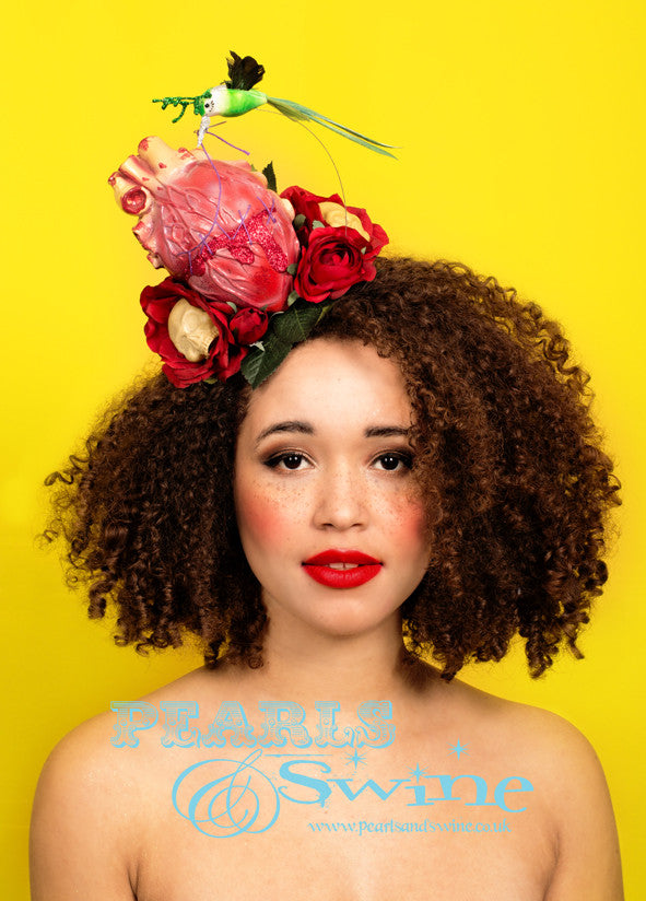 Surreal anatomical heart bleeding red glitter. A hummingbird suspended on wire, with green glitter deer antlers, is stitching the broken heart up with a needle and thread, the bird moves when you wear this. The fascinator base is covered in leaves and red roses with skulls.This limited edition fascinator is inspired by Glenn Arthur's amazing art. The base is backed with leopard print satin and attaches with a comb and hat elastic. This fascinator is piece of wearable art that you can display in a cabinet wh