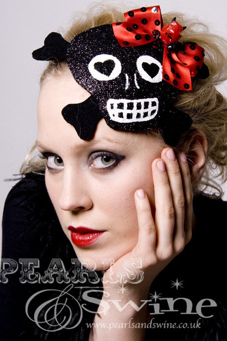 This black glitter skull with cross-bones shaped fascinator decorated with a red bow with devil head and sequins Backed with leopard-print satin, the black glitter skull fascinator attaches with a comb and adjustable hat elastic.