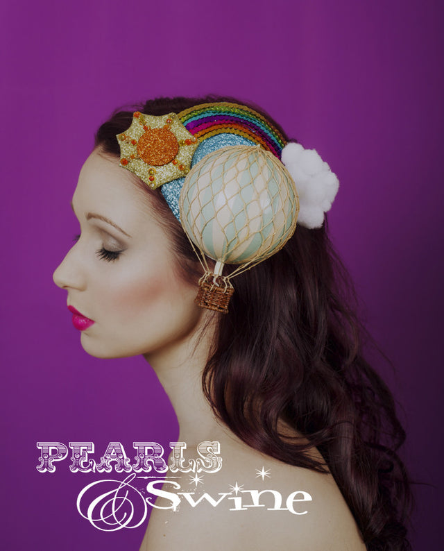 Miniature hot air balloon set on blue glitter sky fascinator base decorated with a sequin rainbow, sun and pom-pom clouds. Backed with satin, this unusual fascinator attaches with a comb and hat elastic. This is a quirky headpiece inspired by summer, it's perfect for a tea party or wedding. It's very sparkly too!