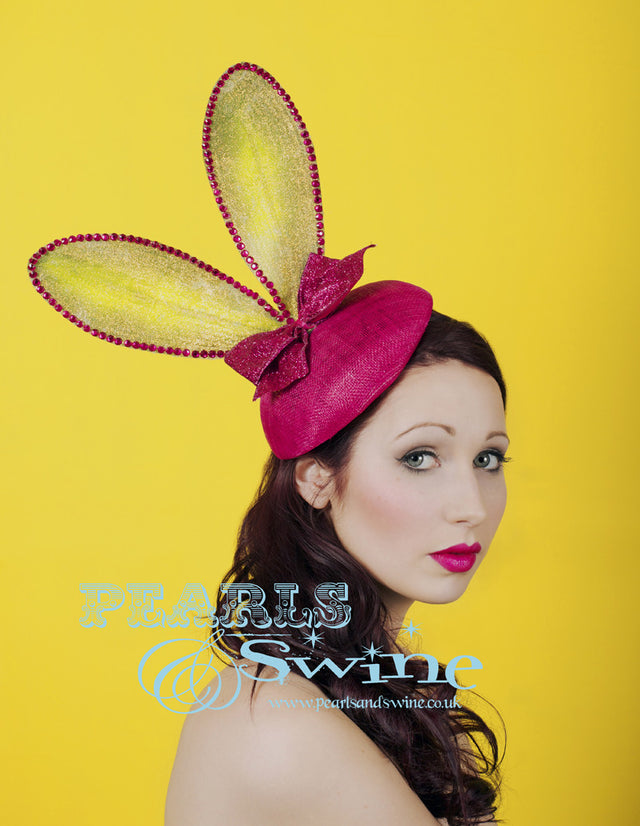 Lime green bunny ears embellished in iridescent gold glitter set on a fuchsia pink sinamay button hat base decorated with a pink glitter bow. This hat attaches with a comb and hat elastic.  This quirky headpiece would be completely at home at Royal Ascot Ladies Day or a Mad Hatters tea party.