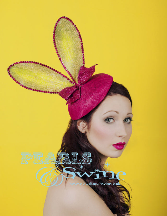 Lime green bunny ears embellished in iridescent gold glitter set on a fuchsia pink sinamay button hat base decorated with a pink glitter bow. This hat attaches with a comb and hat elastic. This quirky headpiece would be completely at home at Royal Ascot Ladies Day or a Mad Hatters tea party.