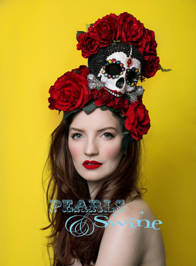A Frida Kahlo inspired Day of the Dead sugar skull hat. The glittered sugar skull is decorated with long 3D eyelashes, it is hand-painted, decorated with crystals. The skull is sat on a silver glittered bone covered in clear diamante gems. The red silk roses are sprinkled with red crystals set on a molded wired sinamay base backed in satin that curves to fit against your head smoothly and the base is covered in red silk roses and leaves. The hat base is backed with leopard print satin and attaches with a co