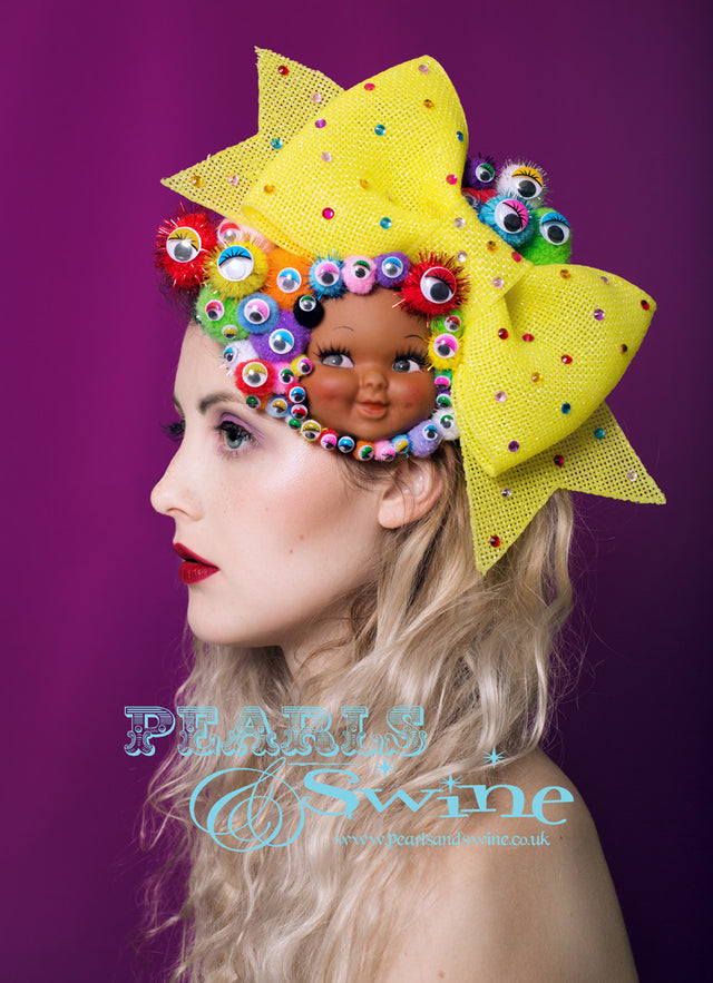 Vintage dimpled doll face fascinator decorated with colourful pompoms, wiggly eyes, a giant yellow glitter bow decorated with gems. All set on a fascinator base which attaches with a comb and adjustable hat elastic. This surreal doll face headpiece is totally eye catching and quirky. Perfect for Royal Ascot Ladies day or a unique bride who wants to make a real statement on her wedding day. This would be fabulous displayed as a piece of art when not worn.
