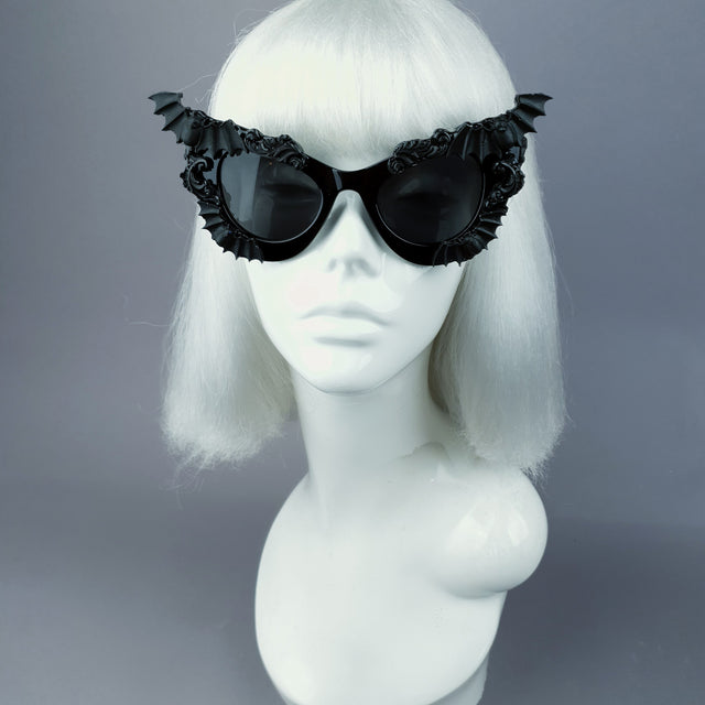 Black cat eye shaped sunglasses decorated with bat wings and filigree, displayed on a mannequin head