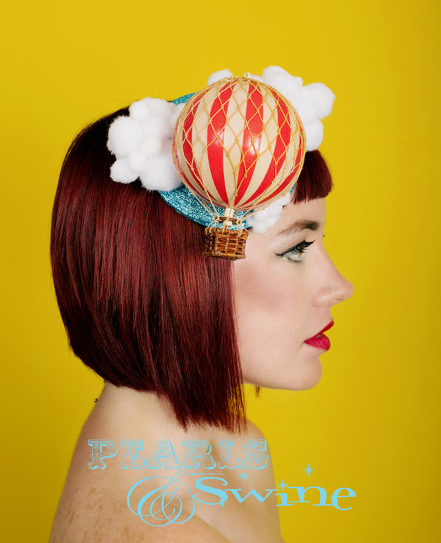 Red and white hot air balloon fascinator, featuring a turquoise blue glitter fascinator sky, with fluffy white pom-pom clouds. The base is backed with leopard print satin and attaches with a comb and adjustable hat elastic. The perfect fascinator for those with their heads in the clouds! It's ideal for a wedding or tea party.