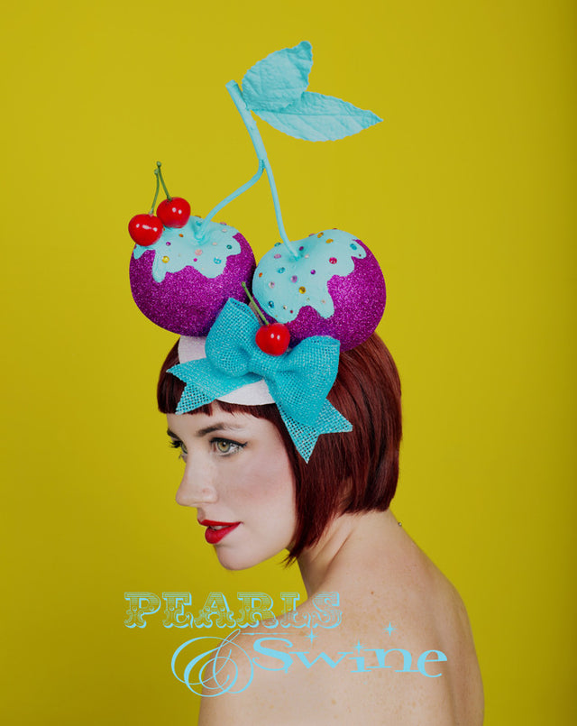 Fuchsia pink glittered giant cherries, hand painted blue "icing" sprinkled with multi-coloured jewel 100s & 1000s, set on an iridescent fascinator base decorated with a blue glitter bow and ruby-red mini cherries. The base is backed with leopard print satin and attaches with a comb and adjustable hat elastic. Perfect for Royal Ascot Ladies Day, giant cherry fans and lady who bake!
