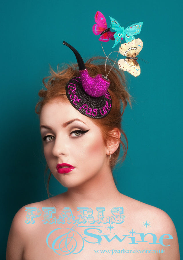 Tribute to "Ceci n'est pas une pipe" by René Magritte, a Belgian surrealist artist aka "This is not a pipe". Nope, it's a fascinator...  Surreal lack fascinator base decorated with a pink glittered pipe, silver glittered wires with colourful butterflies (which come to life and move when you wear this) and the words "Ceci n'est pas une pipe" in pink glitter. This headpiece is backed in leopard print satin and attaches with a comb and adjustable hat elastic.  Perfect for fans of René Magritte's art.