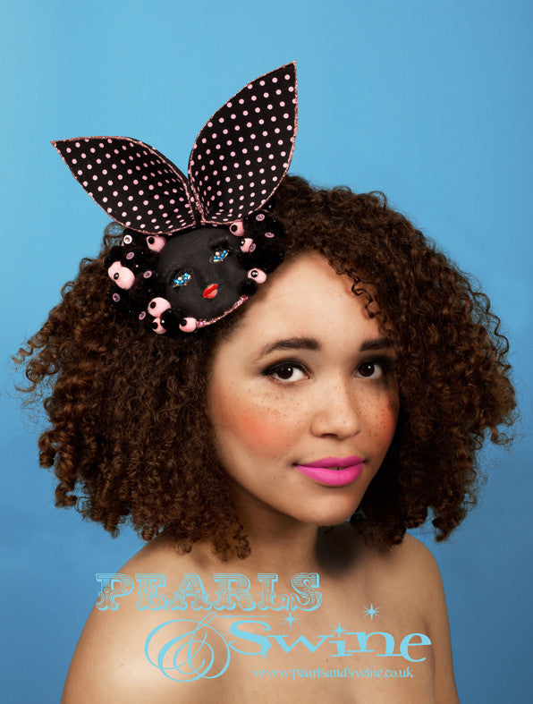 Black vintage doll face fascinator decorated with crystals, pompoms, 3D eyelashes and pink & black polka dot bunny ears edged in a pale pink glitter. Set on a fascinator base this headpiece attaches with a comb and adjustable hat elastic. This whimsical fascinator perfect for Ladies Day at Royal Ascot, it would also make a unique bridal headpiece for an avant garde bride. Suitable for display as a piece of wearable art.