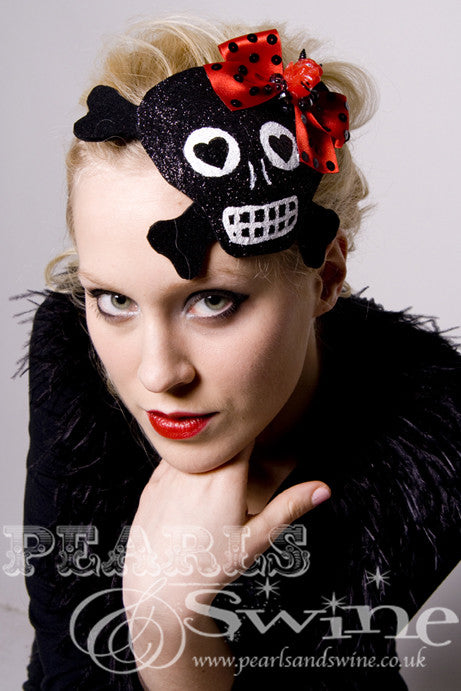 This black glitter skull with cross-bones shaped fascinator decorated with a red bow with devil head and sequins Backed with leopard-print satin, the black glitter skull fascinator attaches with a comb and adjustable hat elastic.