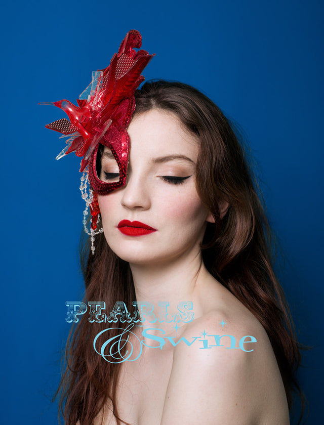 A hand blocked half mask fascinator covered in red glitter, hand stitched pieces of red and white car light, sequin, beading, crystals and a hummingbird bursting out of the explosion. Backed with satin, this attaches with a comb and adjustable hat elastic.