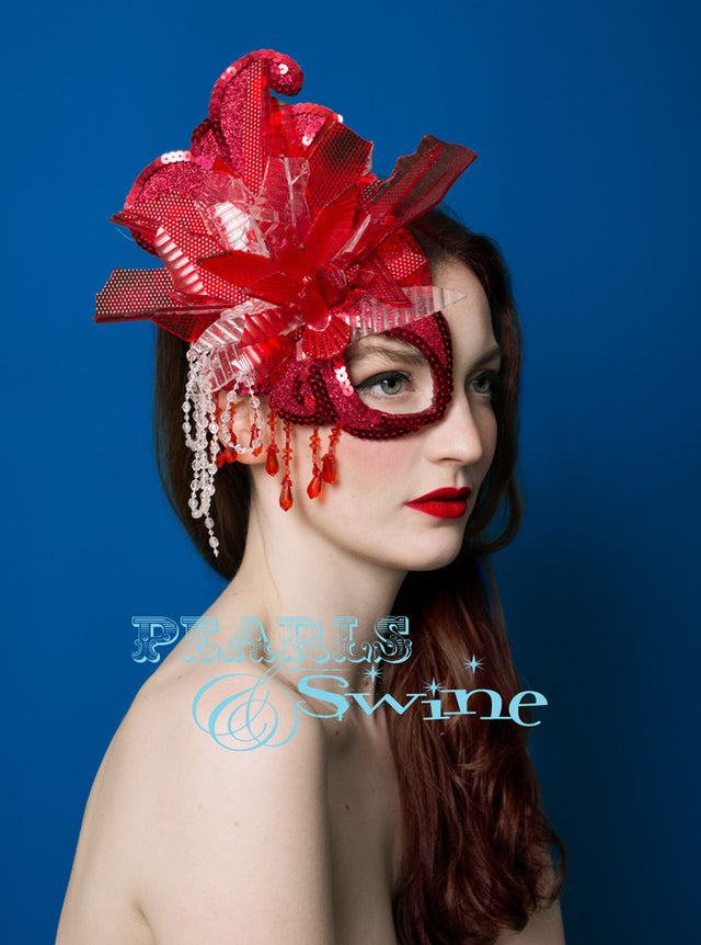 A hand blocked half mask fascinator covered in red glitter, hand stitched pieces of red and white car light, sequin, beading, crystals and a hummingbird bursting out of the explosion. Backed with satin, this attaches with a comb and adjustable hat elastic.