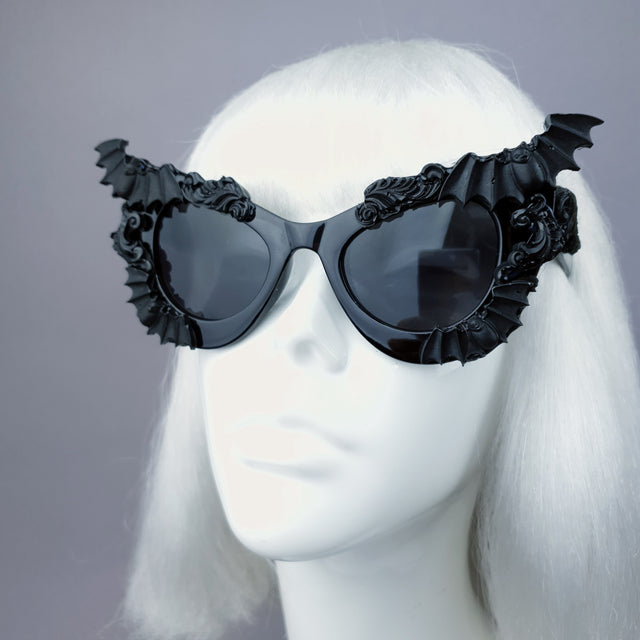 Close up of black cat eye shaped sunglasses decorated with bat wings and filigree, displayed on a mannequin head