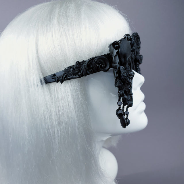Side view of a pair of oversized, ornate black sunglasses with skull, filigree, and bead details displayed on a mannequin head.