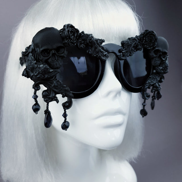 Close-up of a pair of oversized, ornate black sunglasses with skull, filigree, and bead details displayed on a mannequin head.