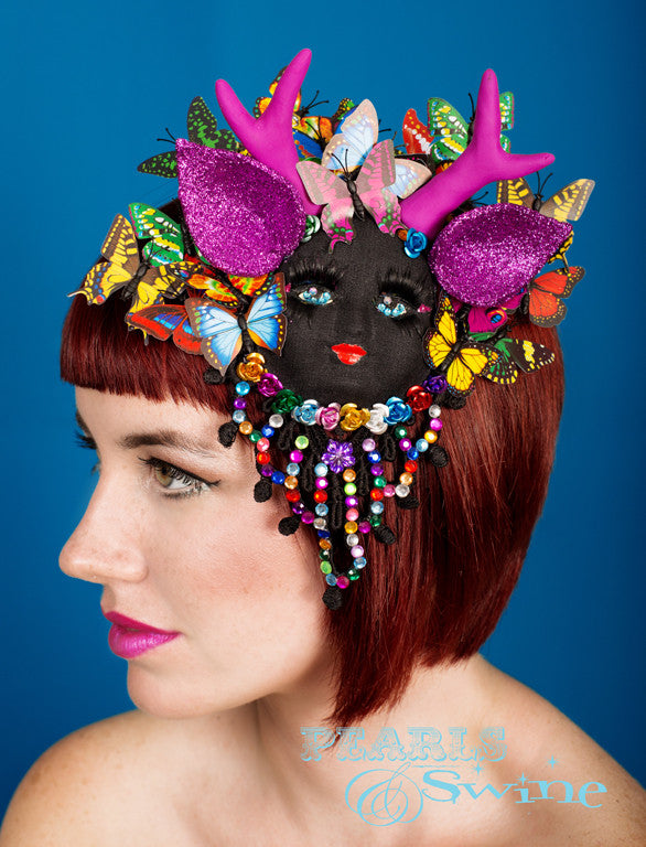 Black vintage doll face antler fascinator with pink glitter ears, pink handmade antlers, long black eyelashes, butterflies and backed with lace covered in multicoloured gems. She is a mythical creature from my imagination, she likes to wear a butterfly headdress, she is a forest spirit. Attaches with a comb and adjustable hat elastic. You can also display this as a piece of art when you aren't wearing it.